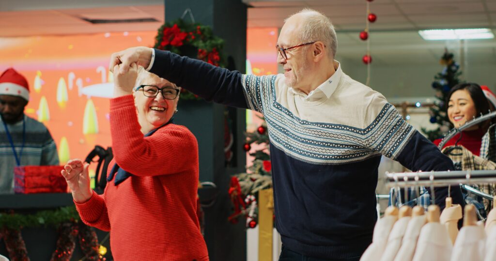 Cheerful elderly couple dancing in Christmas decorated clothing store, feeling xmas holiday mood spirit while browsing for presents. Senior customer being spinned by husband in fashion boutique