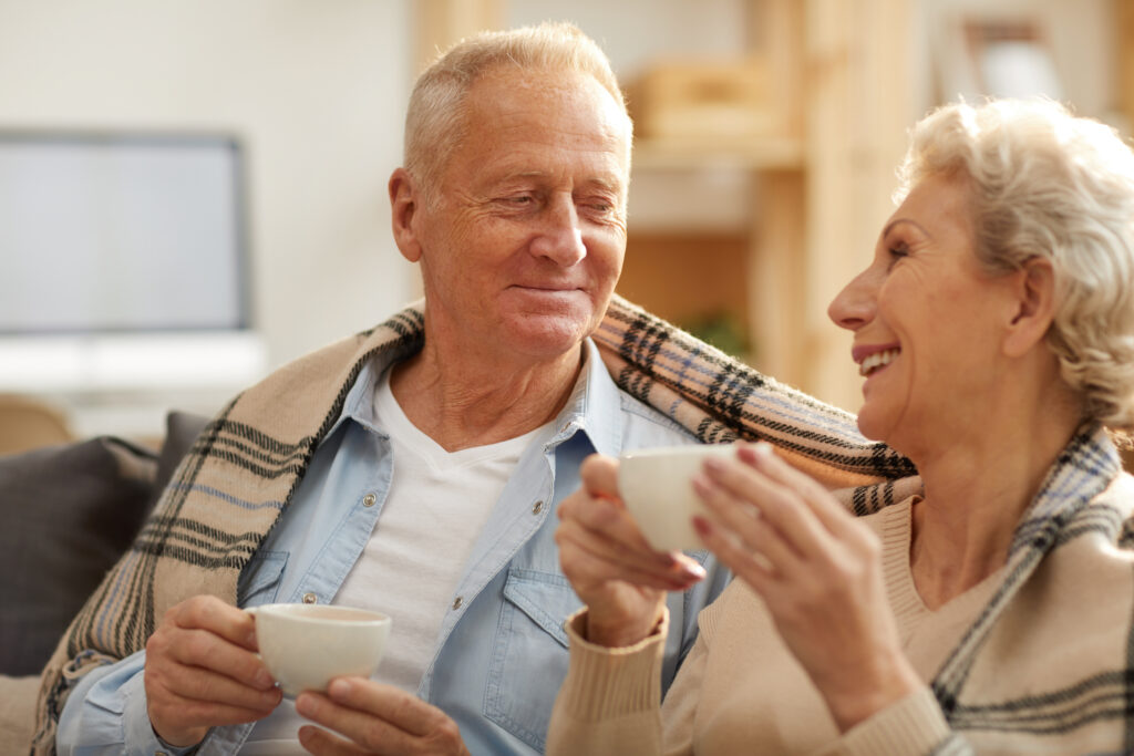 Warm toned portrait of happy senior couple drinking tea sitting on sofa at home lit by sunlight, copy space