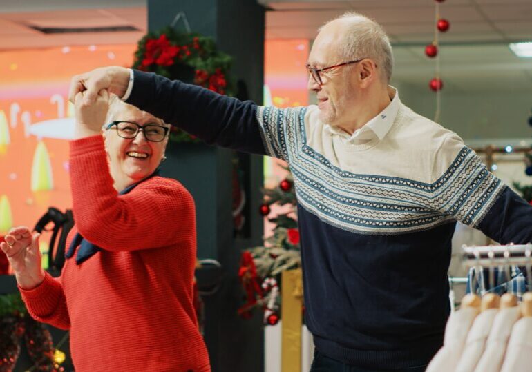 Cheerful elderly couple dancing in Christmas decorated clothing store, feeling xmas holiday mood spirit while browsing for presents. Senior customer being spinned by husband in fashion boutique