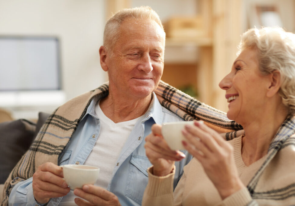 Warm toned portrait of happy senior couple drinking tea sitting on sofa at home lit by sunlight, copy space