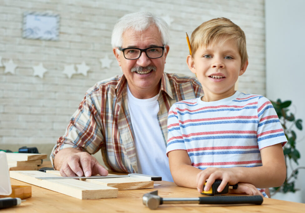 Portrait of grandfather and grandson smiling happily looking at camera while building birdhouse together at home