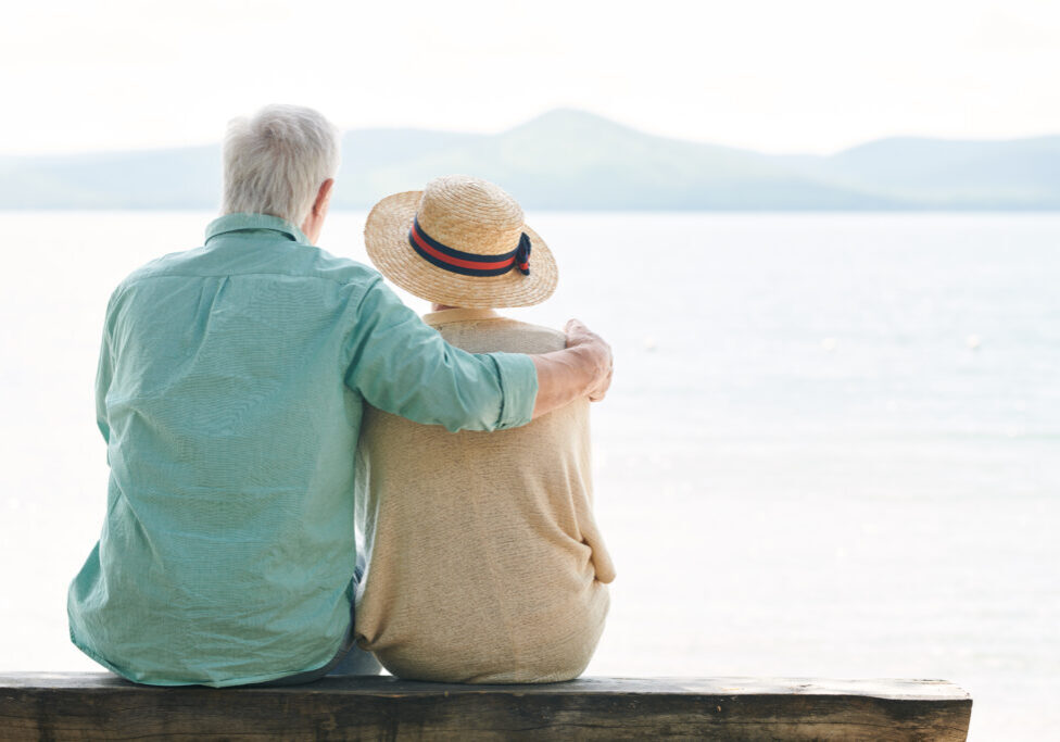 Back view of serene senior couple in casualwear looking at lake while relaxing by waterside on summer day