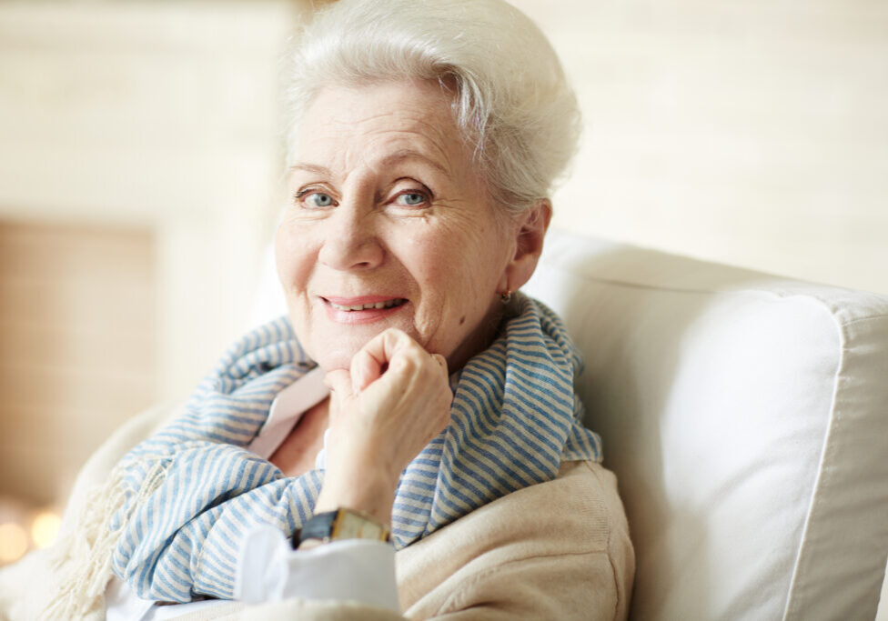 Portrait of beautiful old lady in beige cardigan and striped scarf sitting in armchair, looking at camera and smiling cheerfully